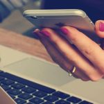 A woman's hands holding an iphone over a laptop on a desk.